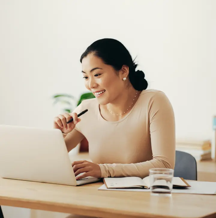 A person sits smiling at their laptop with pen in hand and an open planner next to their elbow and a full glass of water.